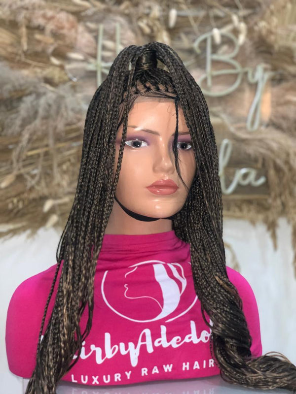 A mannequin head showcases a B29/Ombre French Curls Braided Wig, styled in long, tightly braided hair pulled into a high ponytail. The Ombre French Curls add an elegant touch to the display. Set against a blurred background adorned with decorative elements, the mannequin dons a pink shirt featuring the "Hair by Adedola" logo.