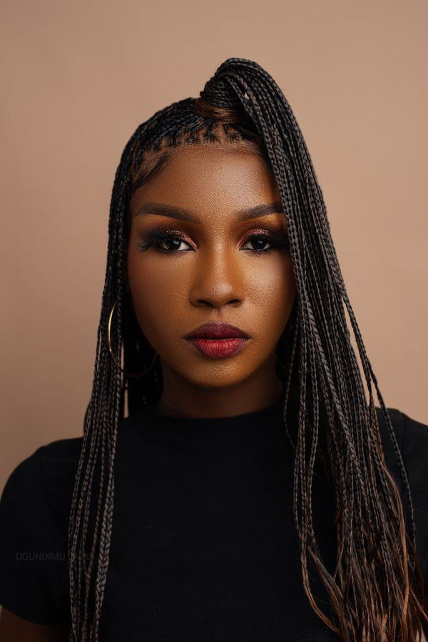 A portrait of a person with long hair styled in the B29/Ombre French Curls Braided Wig by Hair By Adedola, wearing hoop earrings and a black top, set against a plain beige background.